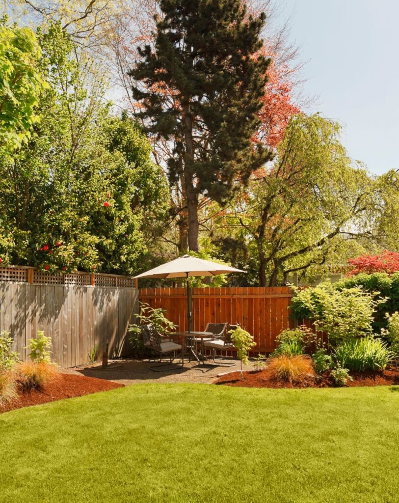 A beautifully maintained backyard with lush green grass, vibrant plants, a wooden fence, and a shaded seating area under an umbrella, illustrating tree care services in Las Vegas.